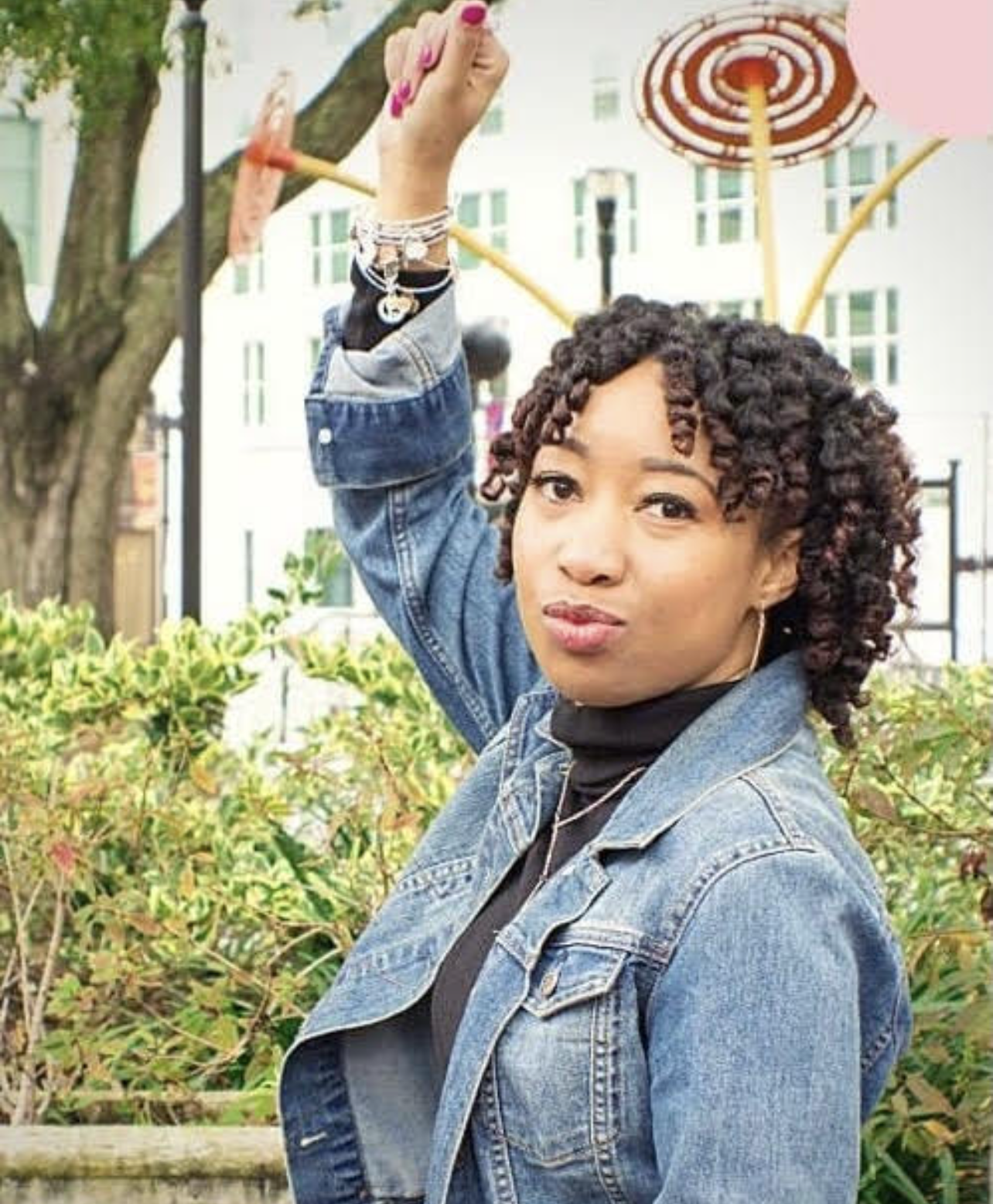 Jessica raises her fist in a confident pose, wearing a denim jacket and black top. She stands outdoors with curly hair, looking boldly into the camera.