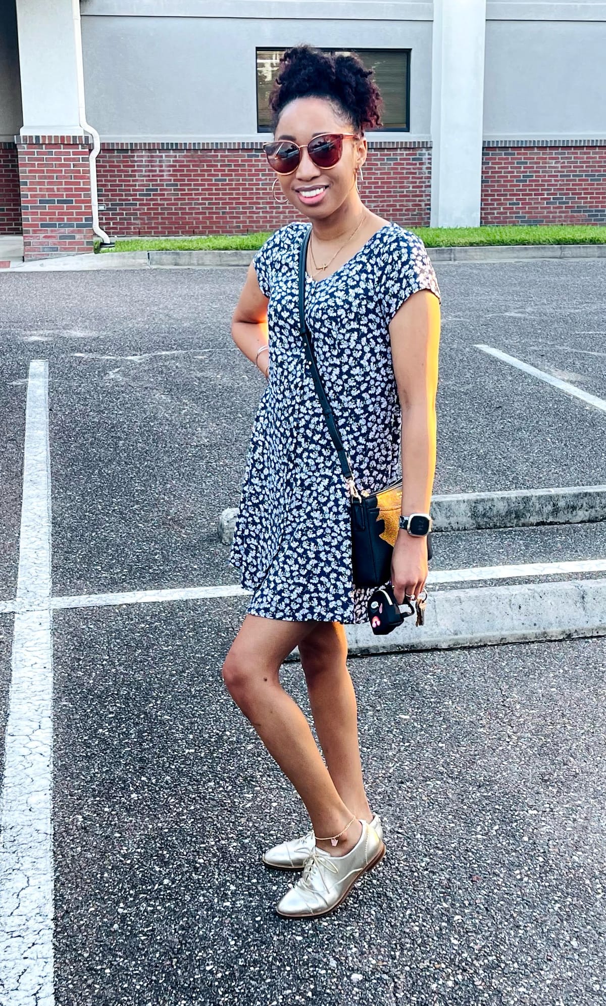 A confident woman smiles in the morning light, wearing a navy floral dress, gold oxfords, and sunglasses. She’s ready for work—stylish, polished, and full of positive energy.