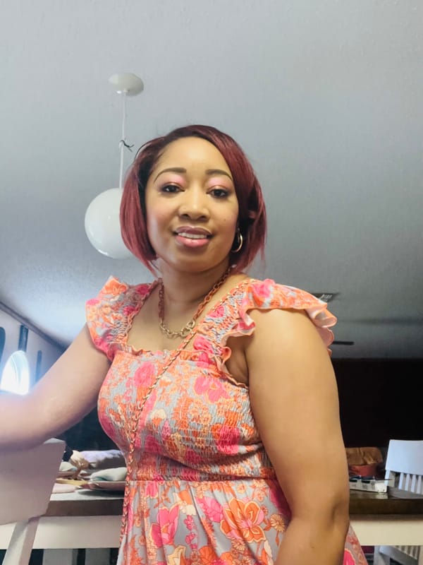 Jessica stands indoors smiling softly. She’s wearing a peach and pink floral dress with ruffled sleeves. Her short hair is styled in a red bob.