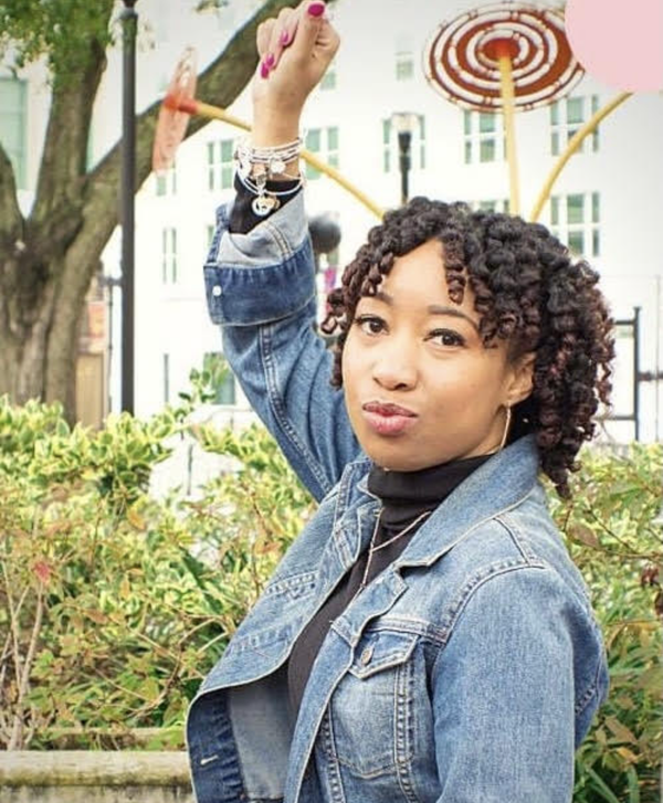 Jessica raises her fist in a confident pose, wearing a denim jacket and black top. She stands outdoors with curly hair, looking boldly into the camera.